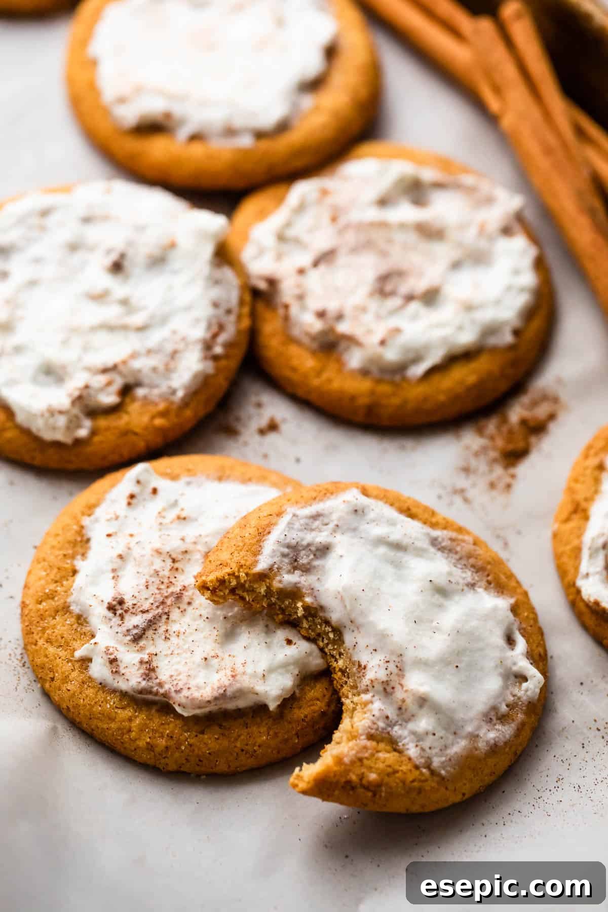 Pillowy Pumpkin Spice Cookies 5 Frosted Pumpkin Cookies arranged on a white background with one cookie halfway bitten off and a few cinnamon sticks set at the top of the right corner.