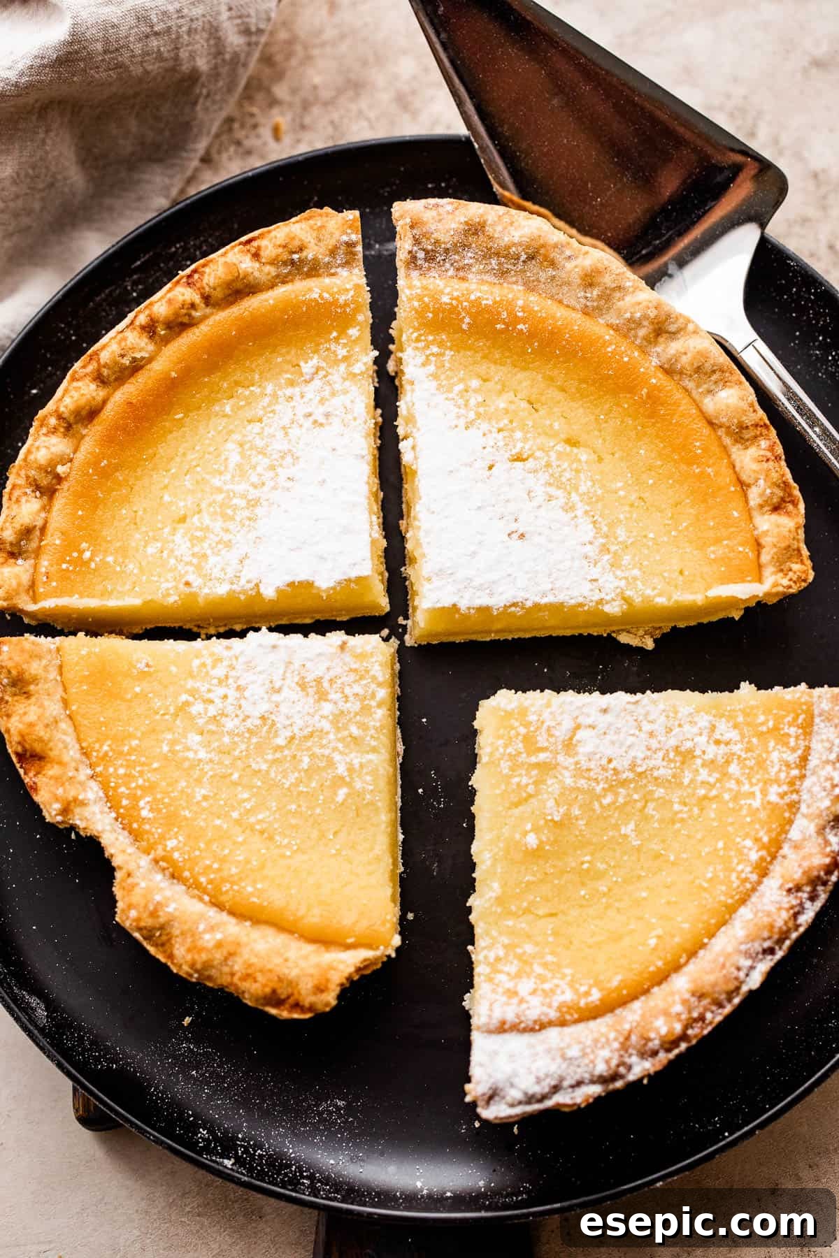 Overhead shot of a black plate displaying four slices of buttermilk pie, each lightly dusted with powdered sugar for an elegant finish.