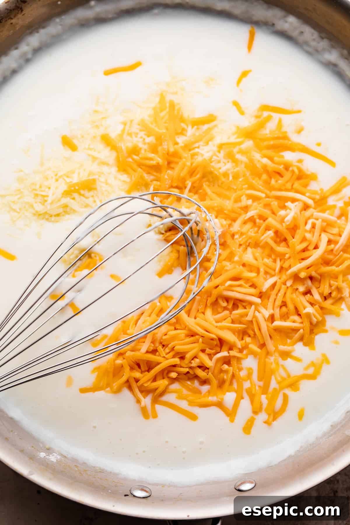 A close-up shot of a whisk vigorously combining shredded cheddar cheese with milk in a large skillet, illustrating the smooth and creamy consistency of the developing cheese sauce.