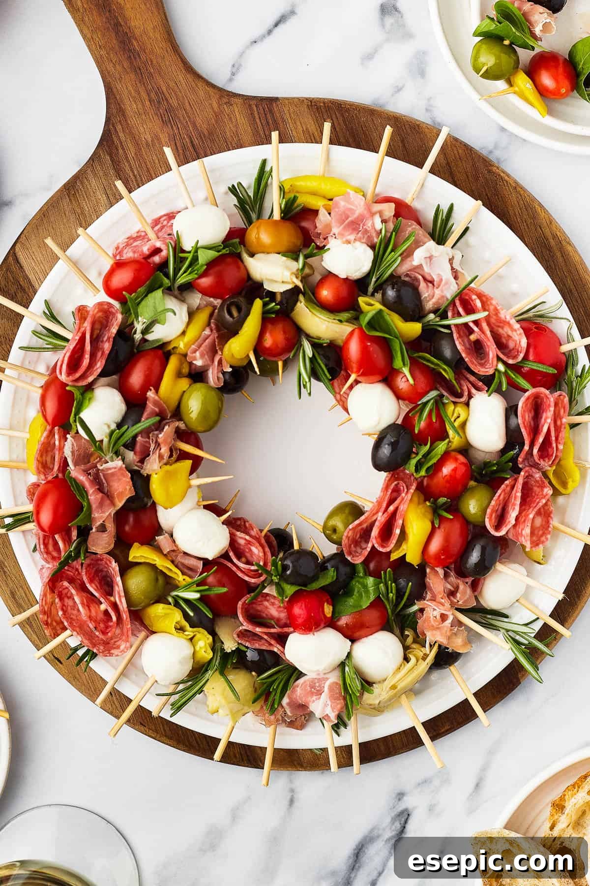 Mini skewers loaded with salami, olives, peppers, and more are stacked and shaped in a ring to make this easy Holiday Antipasto Wreath! A table with a cutting board holding a wreath of skewers, a small pair of scissors, and fresh rosemary sprigs.
