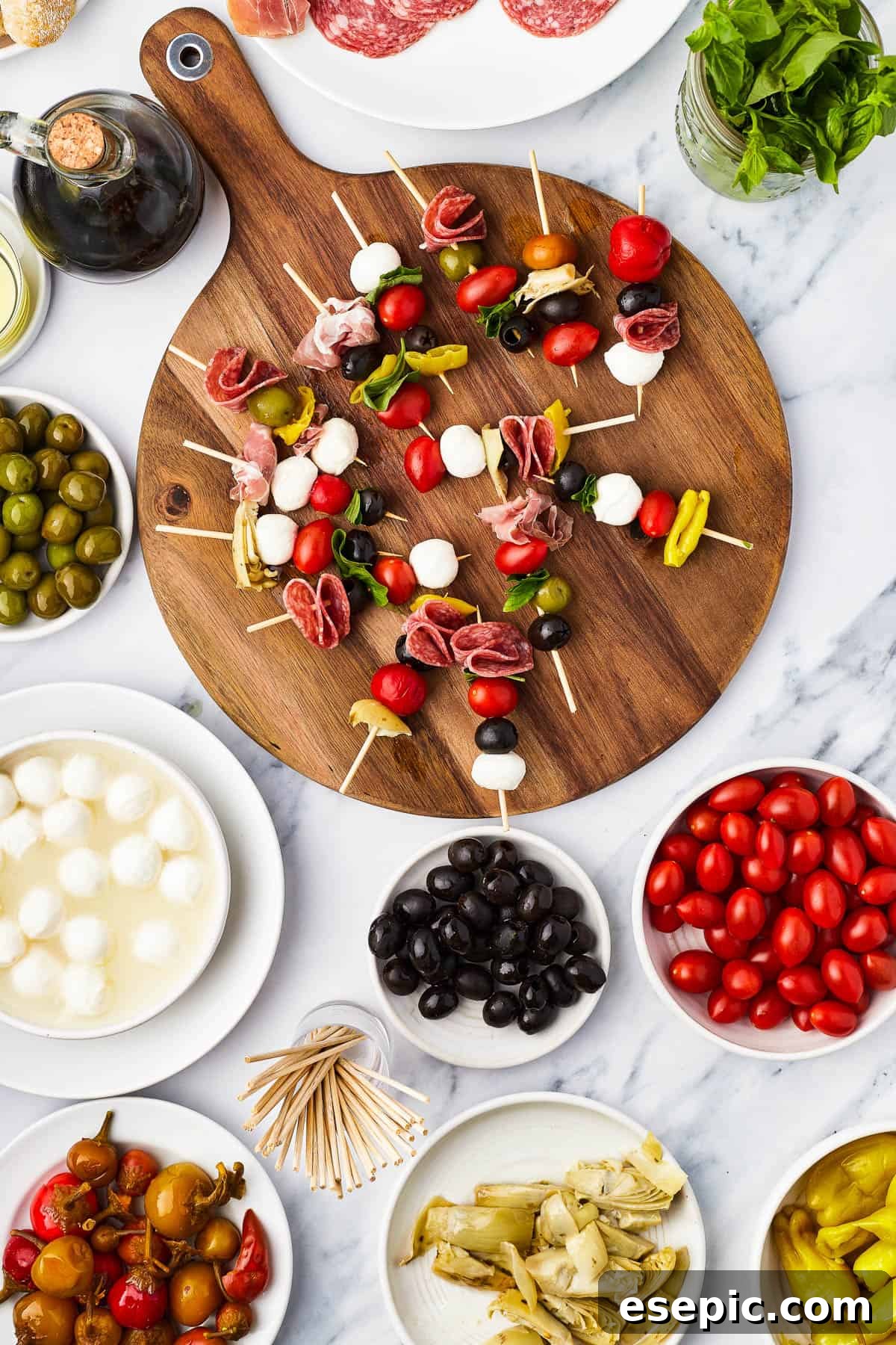 Mini skewers loaded with salami, olives, peppers, and more are stacked and shaped in a ring to make this easy Holiday Antipasto Wreath! Skewers of olives, mozzarella, etc. on a round cutting board, surrounded by bowls of antipasto ingredients.