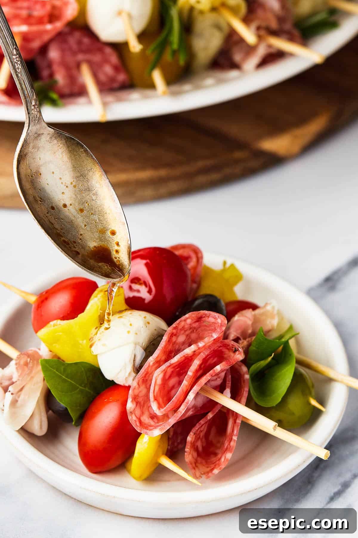 Mini skewers loaded with salami, olives, peppers, and more are stacked and shaped in a ring to make this easy Holiday Antipasto Wreath! A spoon drizzling balsamic vinegar over a serving of antipasto.