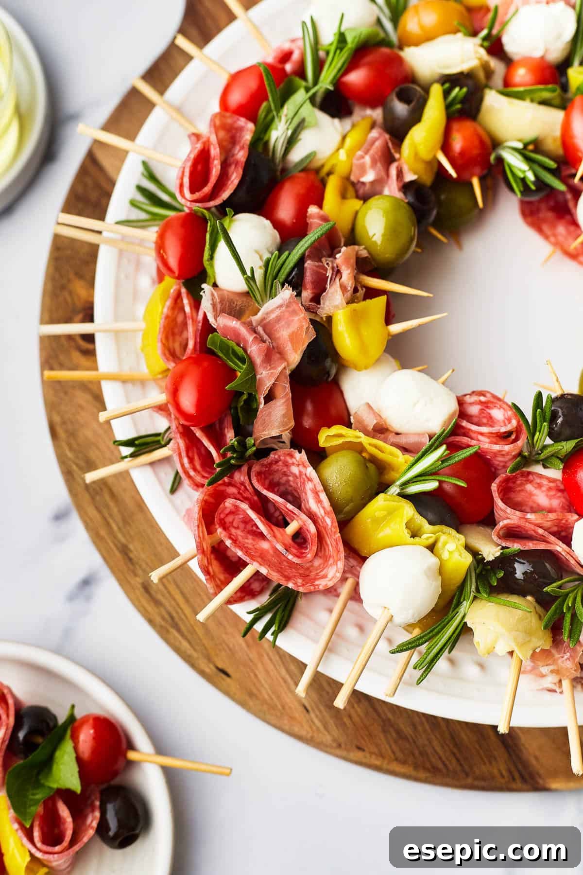 Mini skewers loaded with salami, olives, peppers, and more are stacked and shaped in a ring to make this easy Holiday Antipasto Wreath! A close-up shot of a beautifully arranged holiday antipasto wreath, featuring skewers with prosciutto, pepperoncini, grape tomatoes, mozzarella, and olives, garnished with fresh rosemary.