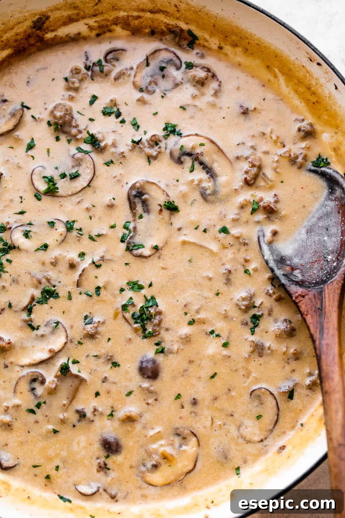 white skillet with simmering beef and mushrooms in a cream sauce with a wooden spoon inside the skillet.
