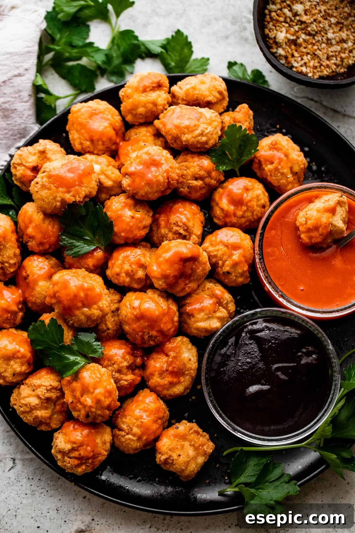 Overhead shot of golden-brown popcorn chicken arranged on a black plate, served alongside small bowls of tangy buffalo sauce and savory barbecue sauce.