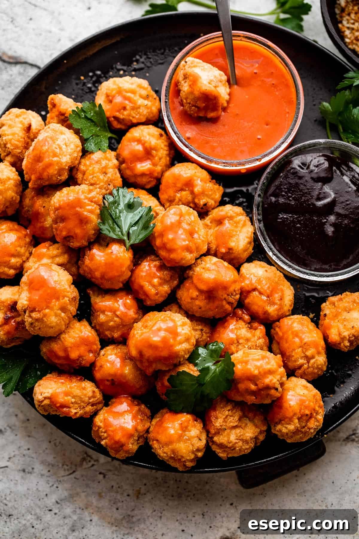 Overhead shot of golden-brown popcorn chicken arranged on a black plate, with bowls of zesty buffalo sauce and sweet barbecue sauce on the side.