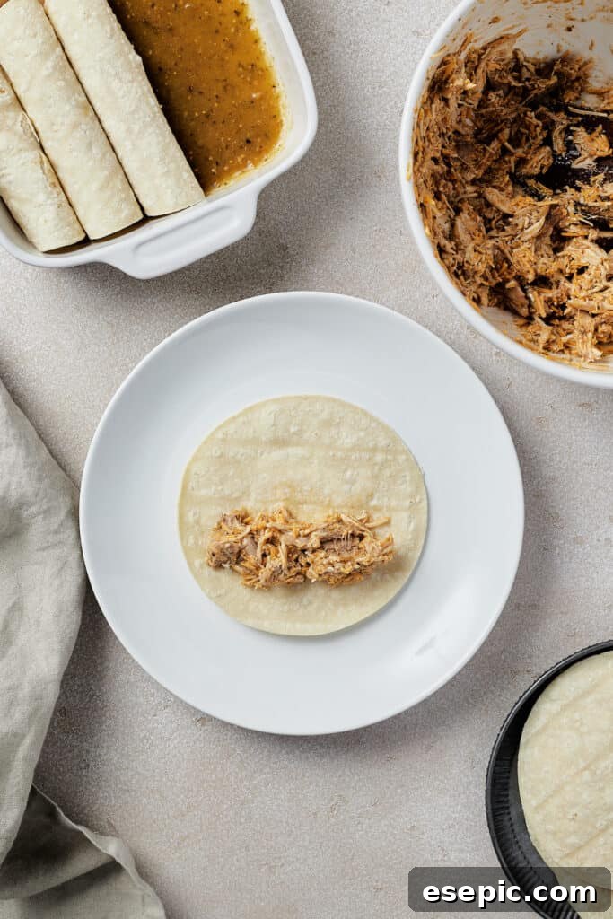 Tangy Tomatillo Chicken Enchiladas 6 A small corn tortilla on a plate with a spoonful of chicken filling arranged on it. A dish of enchiladas, a bowl of filling, and more tortillas are visible in the edges of the frame.