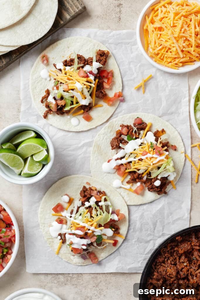 Quick and Flavorful Ground Beef Tacos 6 Tortillas on a work surface. Each tortilla is topped with taco meat and various toppings. A bowl of lime wedges and a small plate of cheese are also on the work surface.