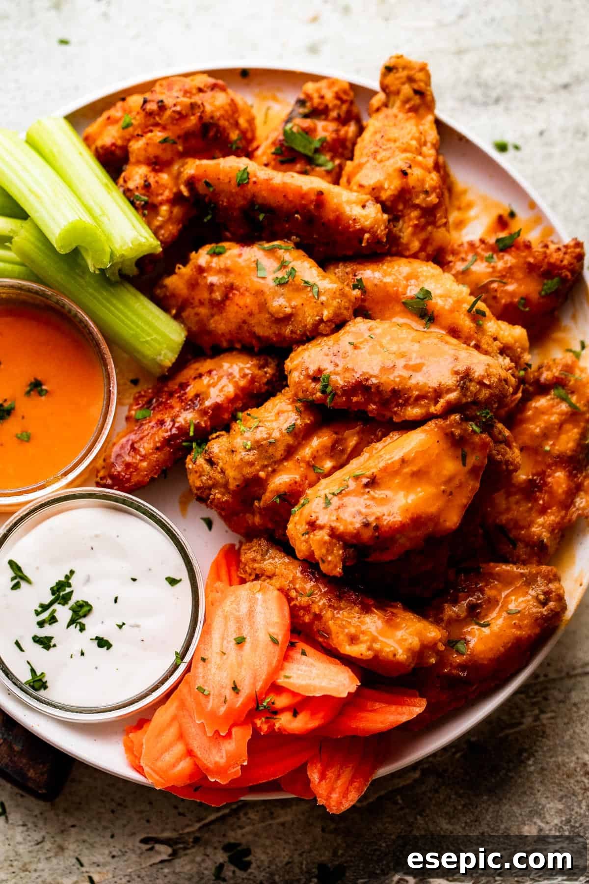 Overhead shot of vibrant Mango Habanero Hot Wings artfully arranged on a pristine white serving plate. Fresh, crisp celery sticks and carrot batons are elegantly positioned alongside the wings, offering a refreshing contrast. Two small jars, one filled with a vibrant hot sauce and the other with a creamy ranch dipping sauce, complete the presentation on the plate.