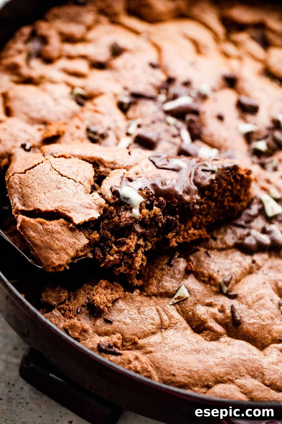 A hand gently pulls a warm, freshly baked wedge of mint chocolate skillet cookie from a cast iron pan, revealing its gooey center and melted chocolate mints.