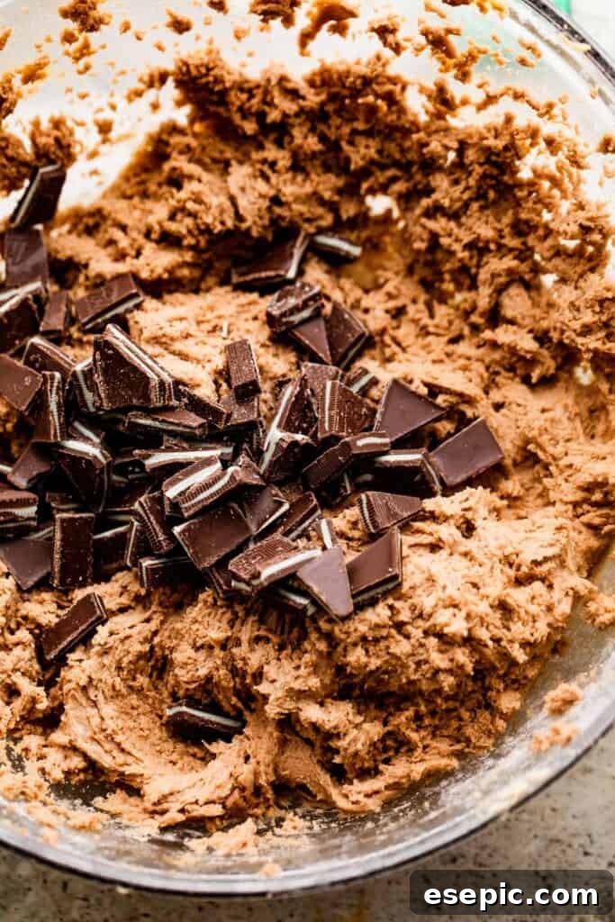 Overhead view of rich cookie dough in a mixing bowl, with a generous amount of chopped chocolate mints scattered across its surface, awaiting incorporation.