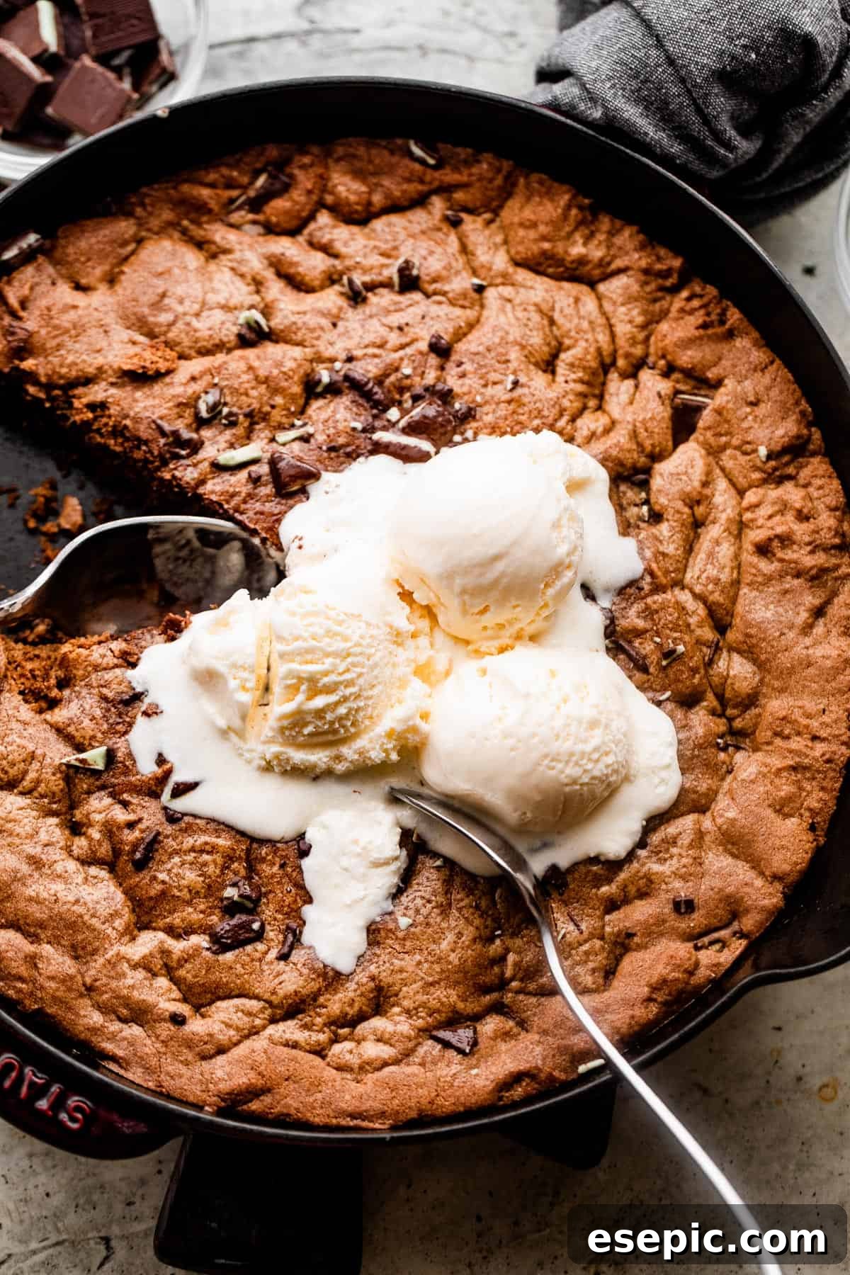 Overhead shot of a delicious mint chocolate skillet cookie still in its pan, topped with three scoops of melting vanilla ice cream and two large spoons ready to serve.