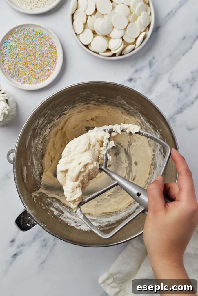 Frosting in a mixing bowl, with a woman's hand lifting a stand mixer attachment covered with frosting, illustrating the smooth texture.