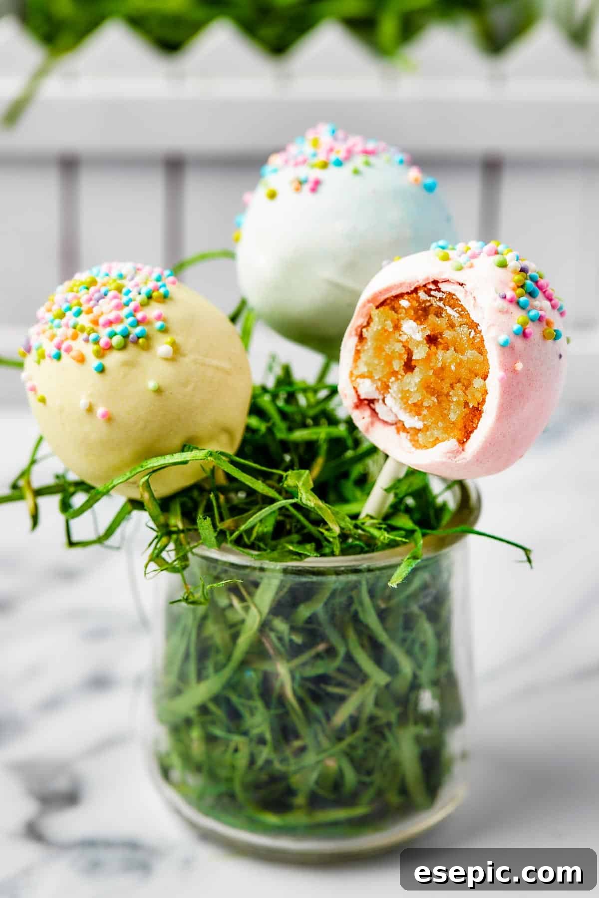 Three pastel cake lollipops arranged in a small glass filled with Easter grass, one with a playful bite taken out.