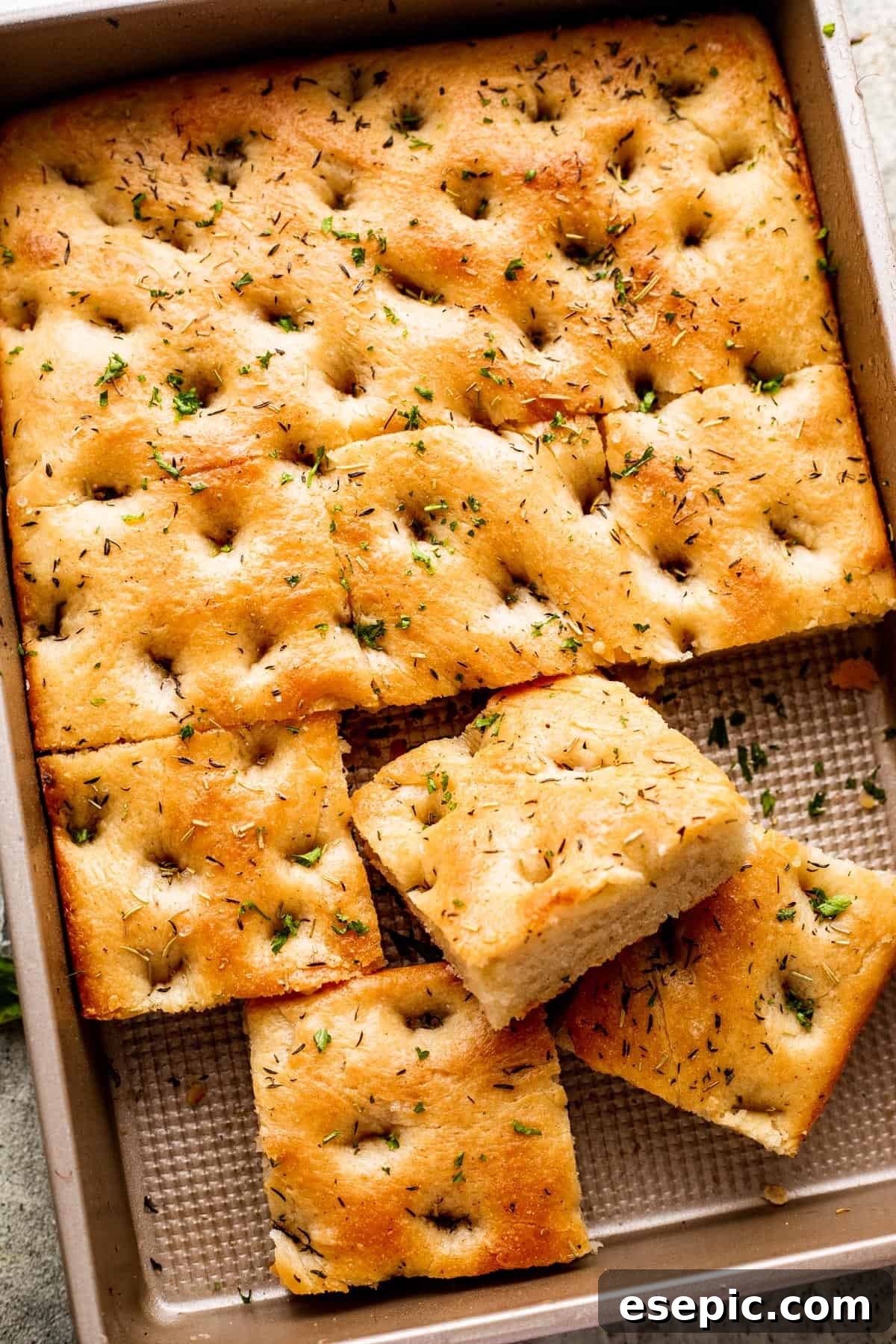 Aromatic Garlic and Herb Focaccia 2 A large pan-sized loaf of focaccia, with four squares of bread cut away, showcasing its golden-brown crust and herb toppings.