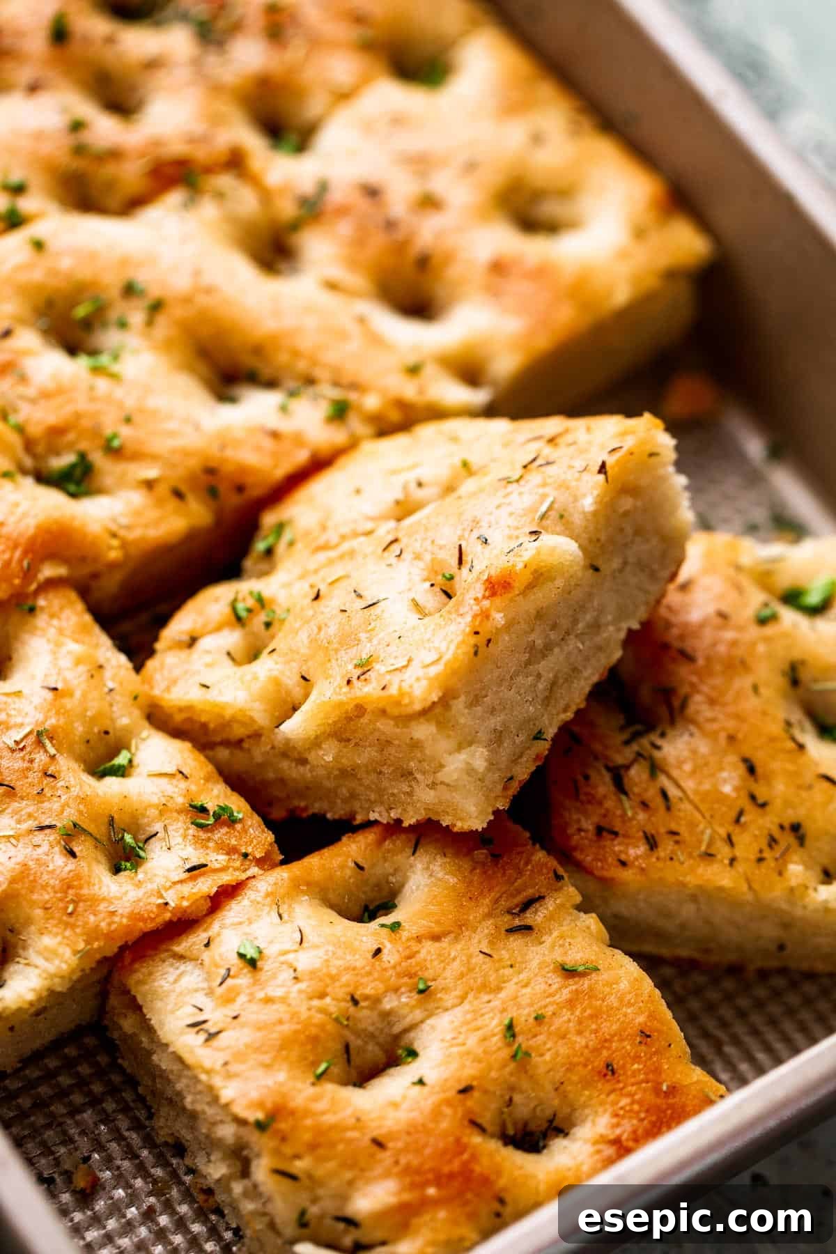 Aromatic Garlic and Herb Focaccia 9 A large pan-sized loaf of focaccia, with four squares cut away, showcasing the airy interior and herb-studded crust.