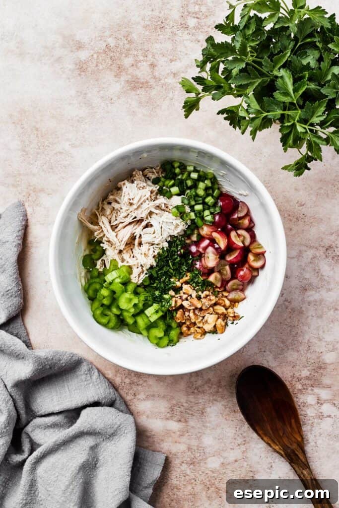 A white mixing bowl with shredded chicken, grapes, celery, and other ingredients, ready for dressing.