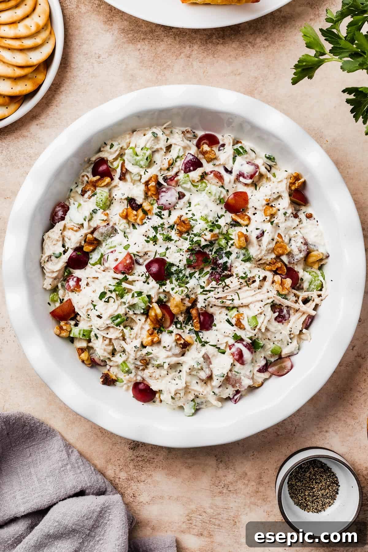 A shallow white dish filled with creamy chicken salad with grapes, garnished with toasted walnuts and fresh parsley.