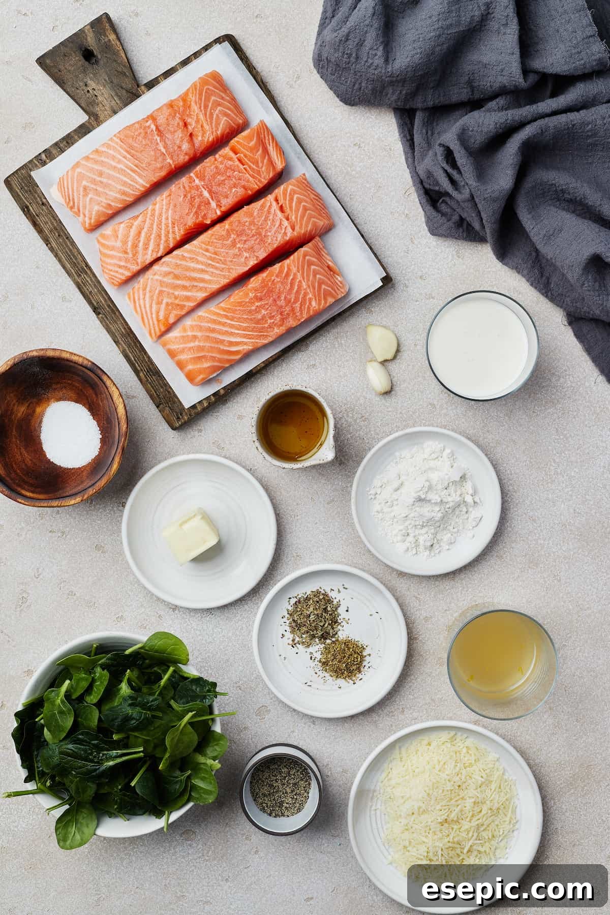 An array of fresh ingredients laid out for Salmon Florentine preparation, including salmon fillets, garlic, heavy cream, salt, butter, olive oil, flour, dried herbs, chicken broth, spinach, pepper, and Parmesan cheese.