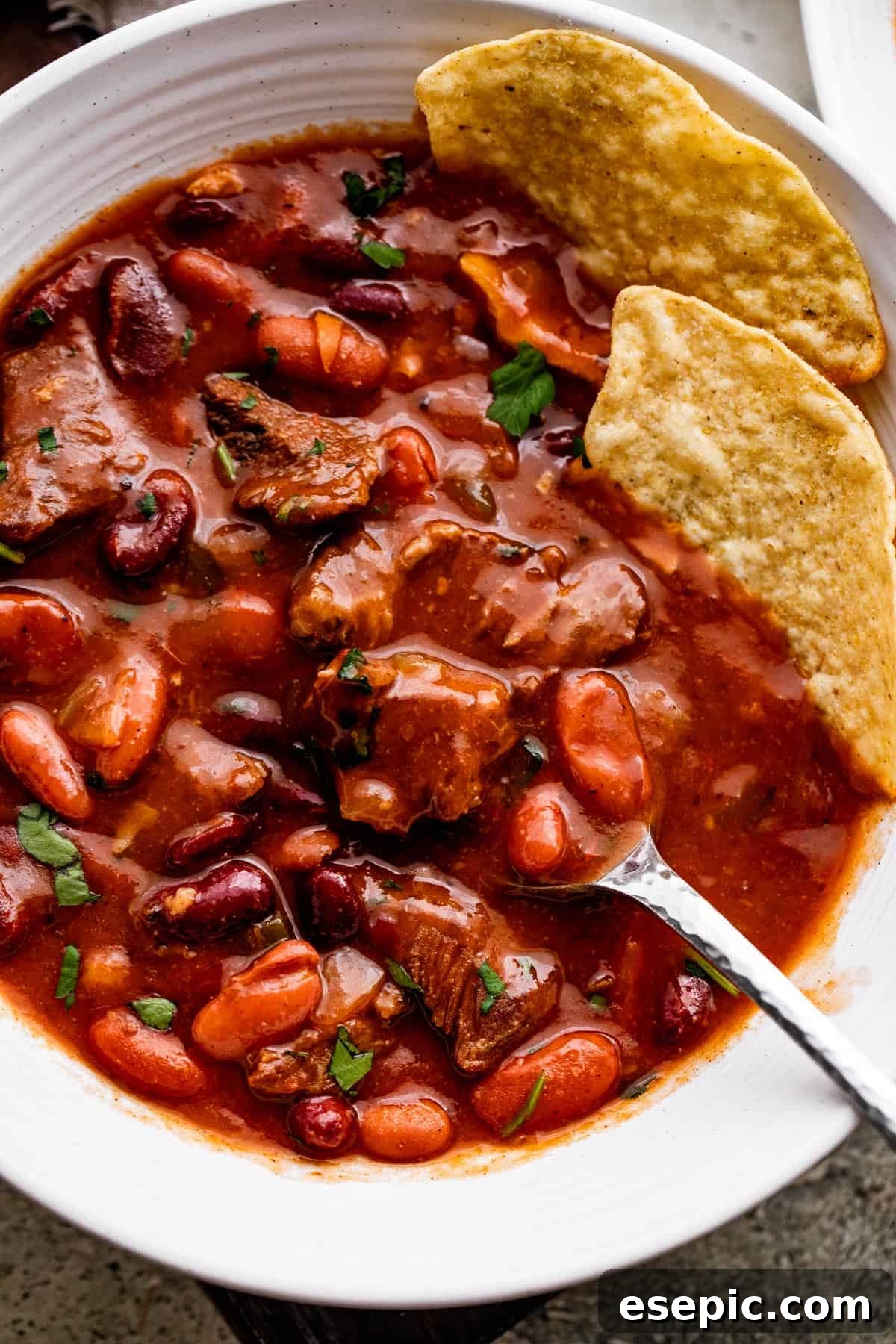 Close-up overhead shot of savory Chili con Carne served in a white bowl, garnished with a spoon and two tortilla chips.
