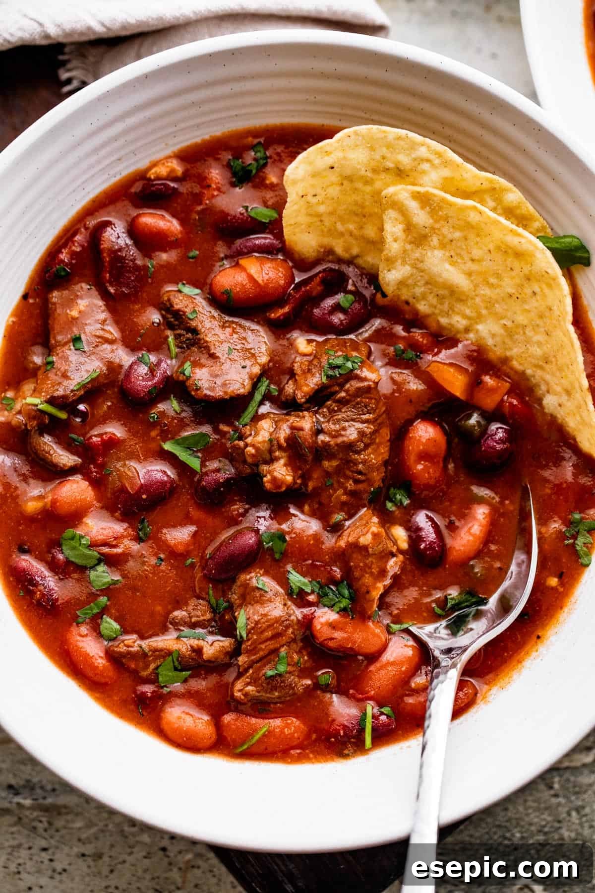 Overhead shot of Chili con Carne in a white bowl, with a spoon and tortilla chips nestled in the chili, ready to be enjoyed.