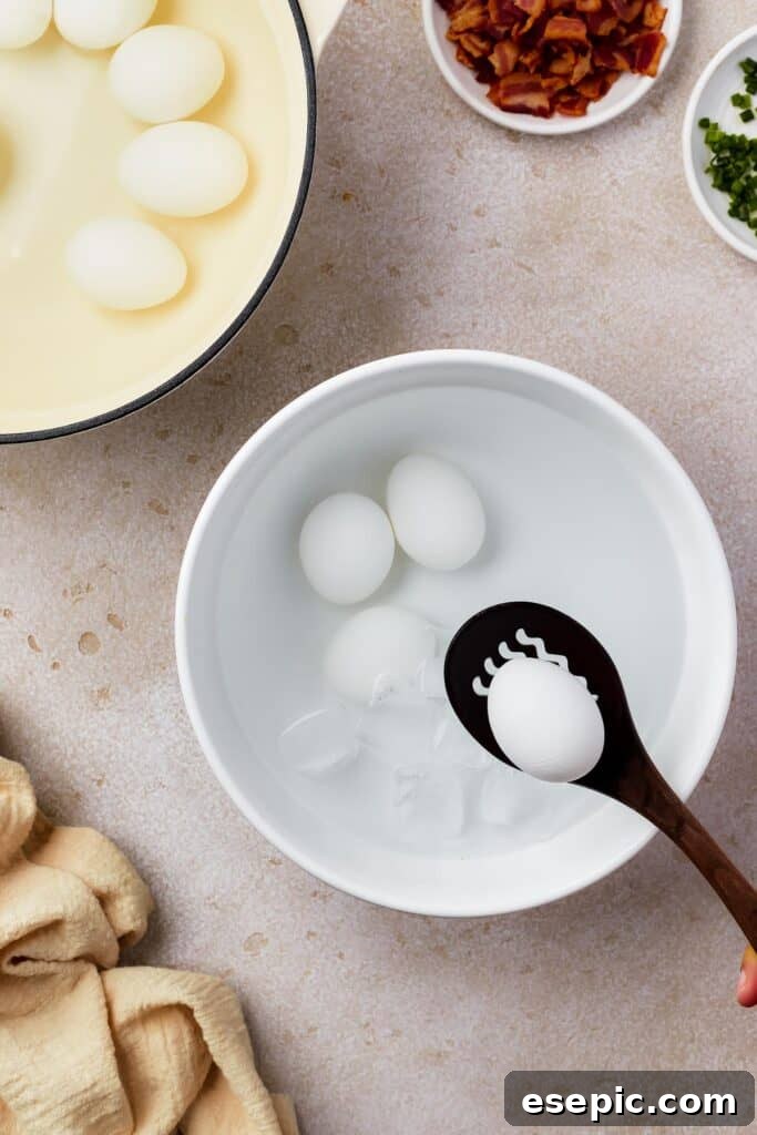 A bowl of peeled hard cooked eggs in water. Nearby are a plate of eggs in the shell, and a small dish of discarded egg shells, along with a few small dishes of ingredients and a cloth napkin.
