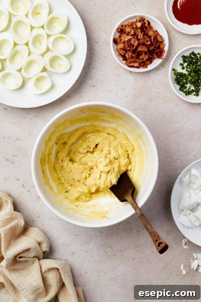 A bowl of creamy egg yolk filling with a wooden spoon. Cooked egg whites, bacon, and other ingredients are on the work surface also.