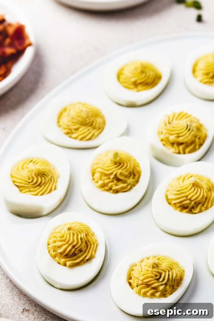 A shallow white platter, shaped like an egg, with deviled eggs arranged on it. Bowls of garnishes are arranged around the platter.