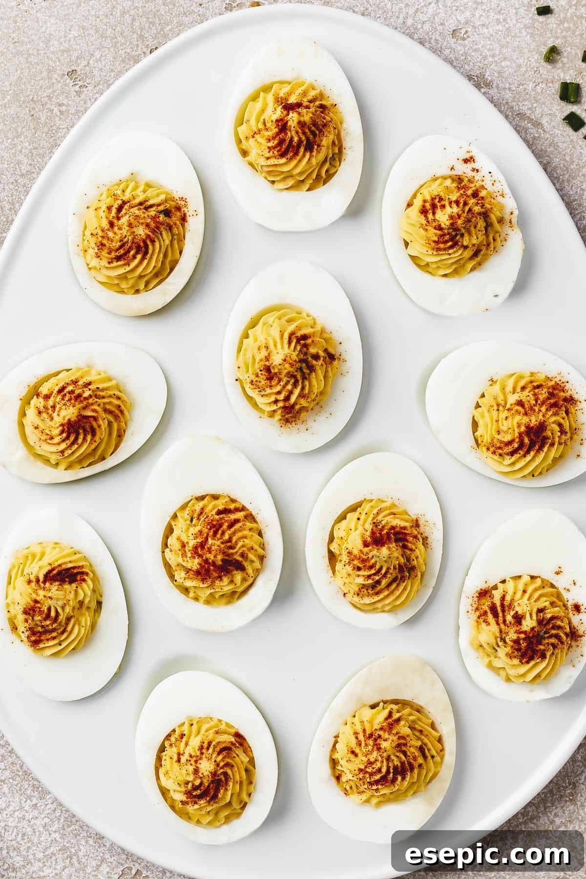 Overhead shot of an egg-shaped white dish with stuffed eggs arranged on it. Each egg has been garnished with paprika, bacon, and chives.