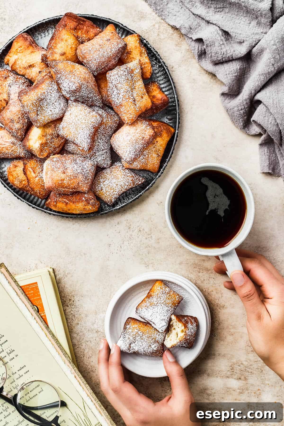 An inviting overhead shot of a large platter overflowing with hot, powdered sugar-dusted beignets. A cup of black coffee sits nearby, completing the classic pairing.