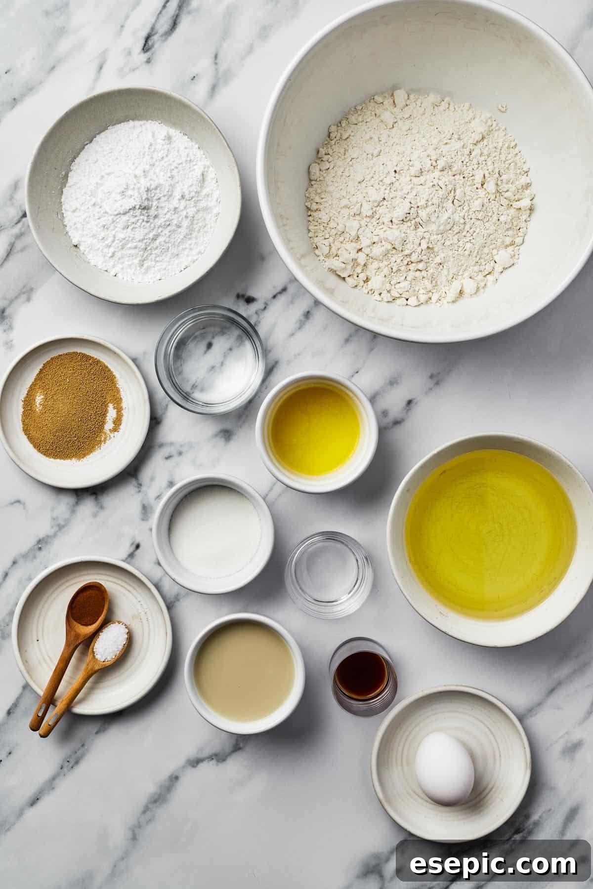 A flat lay photograph showcasing all the essential ingredients for making beignets: powdered sugar, all-purpose flour, active dry yeast, warm water, melted butter, canola oil, granulated sugar, white vinegar, ground cinnamon, kosher salt, vanilla extract, and one egg.