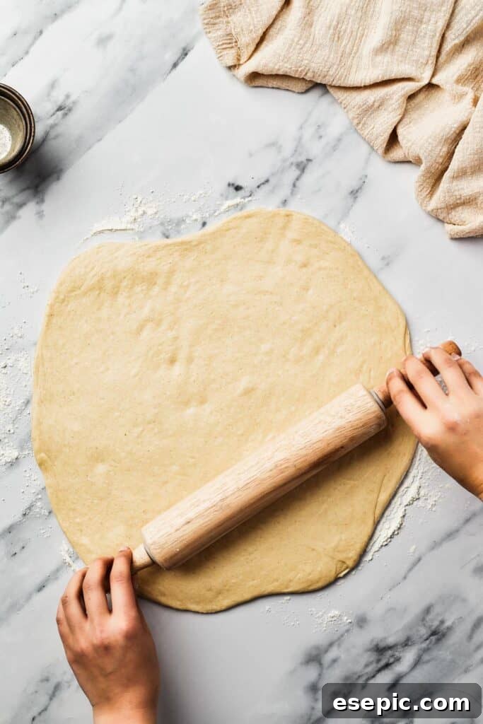 Yeast dough being expertly rolled out into a thin, even layer on a clean marble work surface, preparing for shaping.