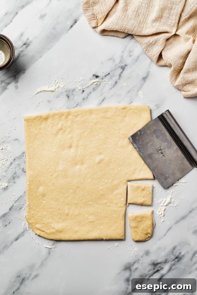 A neatly rolled-out square of yeast dough on a marble work surface, with small, precise square pieces already cut out. A bench scraper rests beside the dough, ready for use.