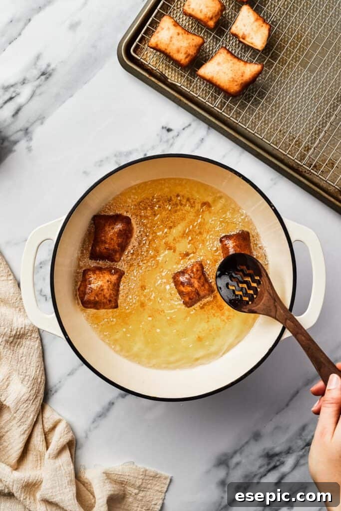Close-up of golden-brown, perfectly fried beignet squares puffing up in a Dutch oven, partially filled with shimmering hot oil.