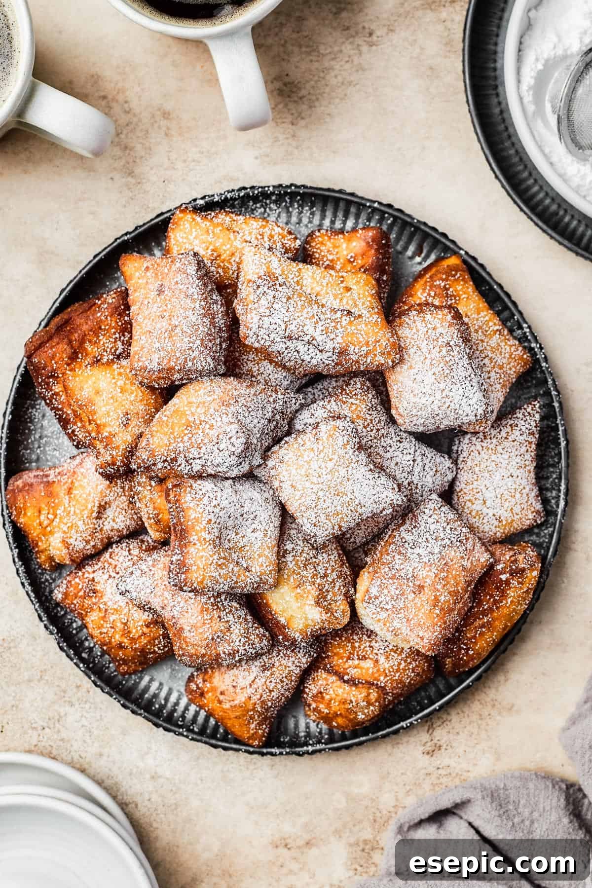 A pristine white plate holds a stack of golden, fried beignets, each generously dusted with a fine layer of powdered sugar.