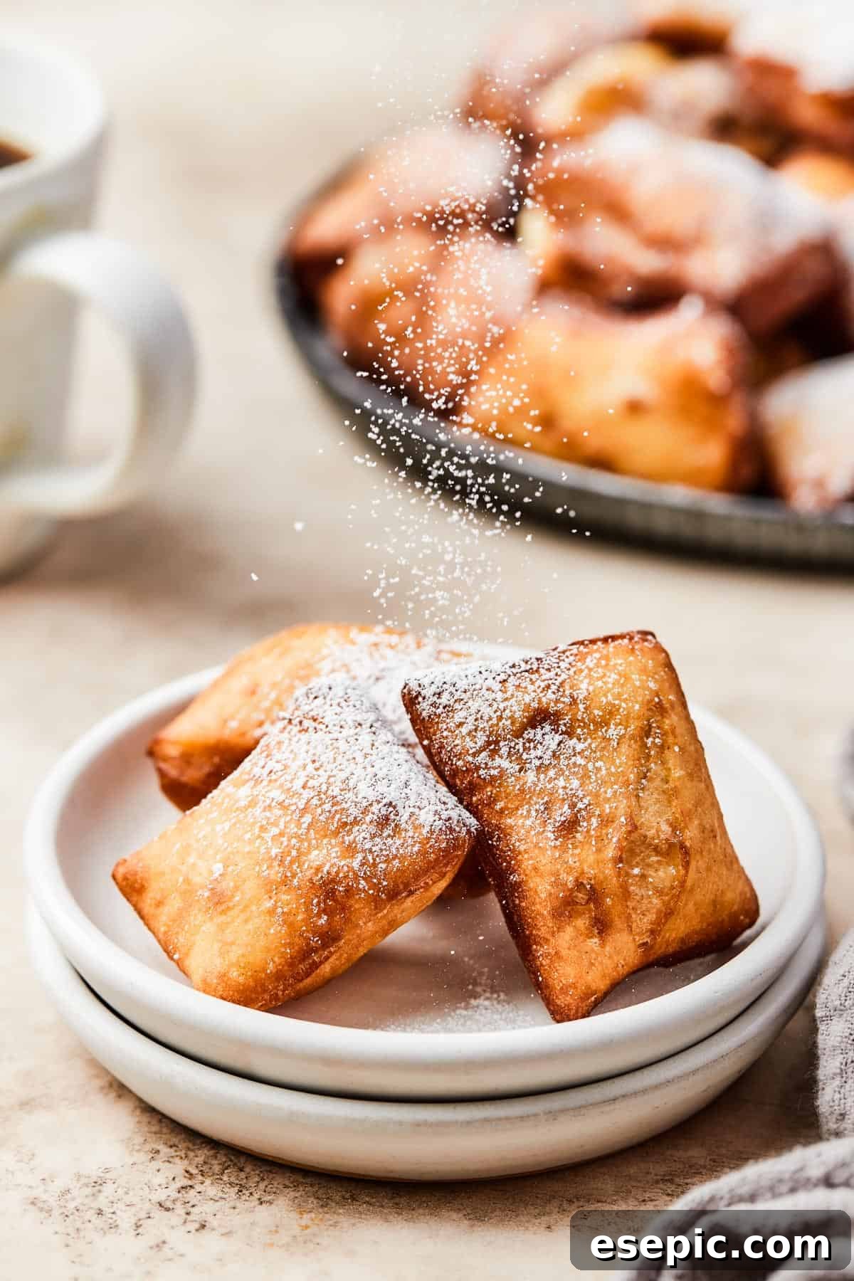 A close-up shot of a hand gracefully sprinkling a generous amount of powdered sugar over three freshly fried beignets, artfully arranged on white plates.