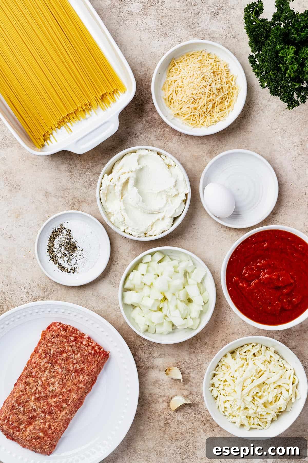 Irresistible Golden Spaghetti Bake 4 A flat lay arrangement of ingredients for baked spaghetti casserole, including dry spaghetti, a chopped onion, various seasonings, ricotta cheese, an egg, mozzarella, marinara sauce, Italian sausage, garlic, and parmesan cheese.