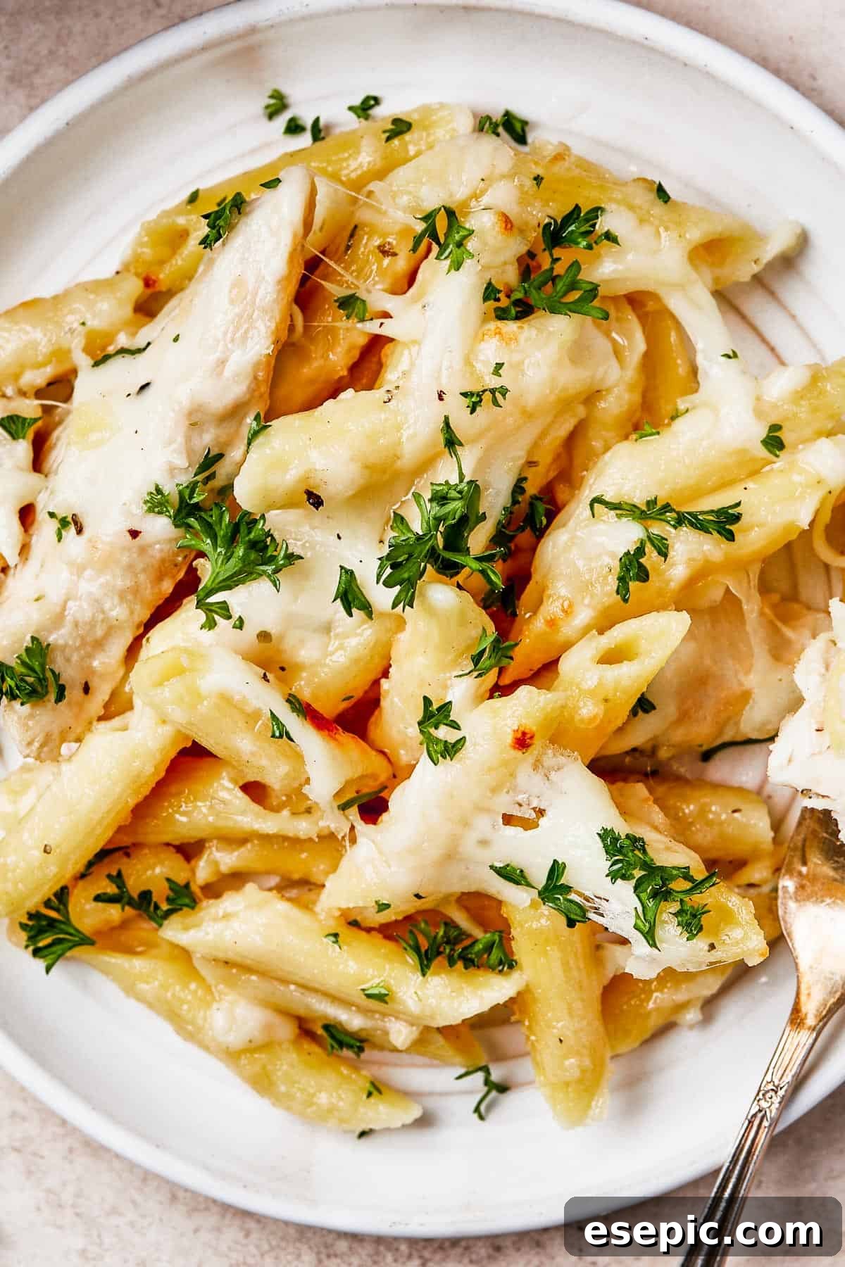 Overhead shot of chicken alfredo pasta bake on a plate, with a fork. The casserole dish is nearby on the table, showing more of the baked pasta ready to be served.