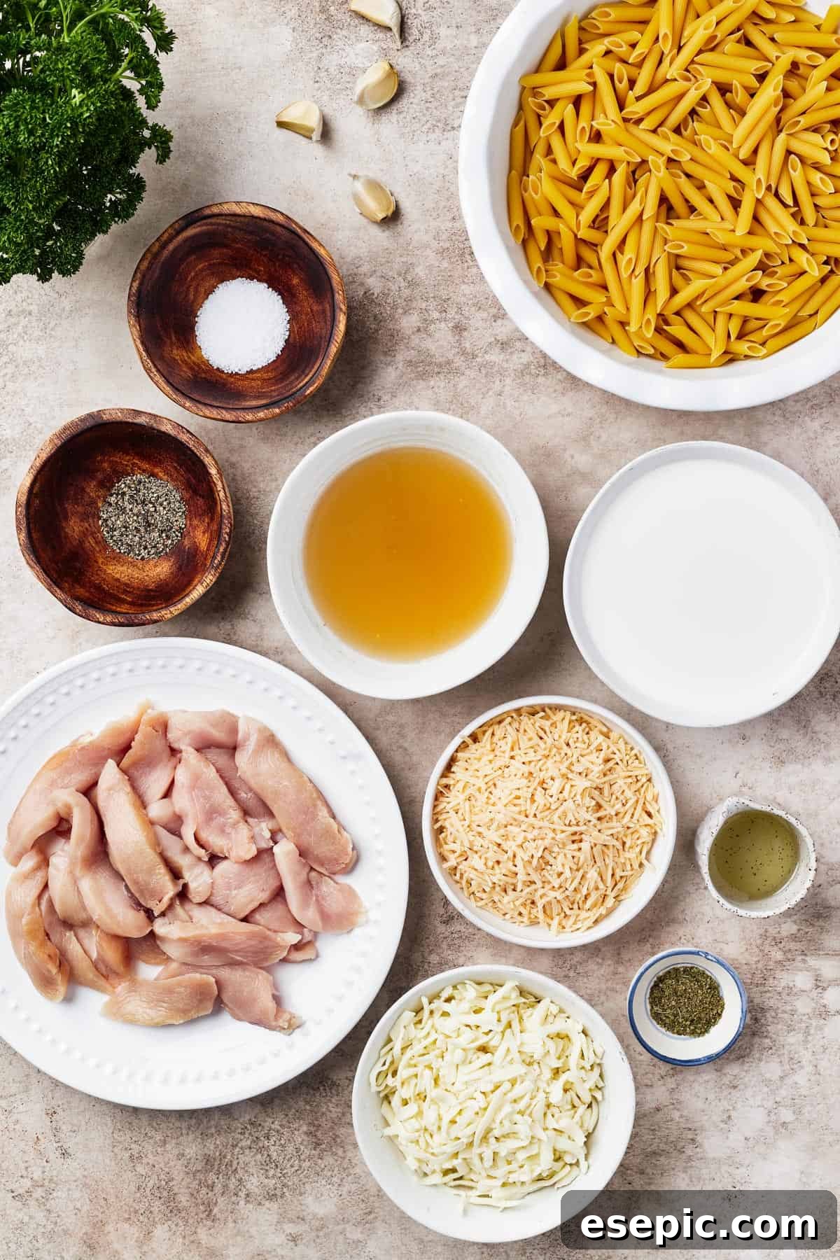 From top left: Fresh parsley, whole garlic cloves, dry penne pasta, salt and ground pepper, chicken broth, heavy cream, raw chicken breast pieces, shredded parmesan cheese, olive oil, shredded mozzarella cheese, and a jar of Italian herbs. These ingredients are neatly arranged on a wooden surface.