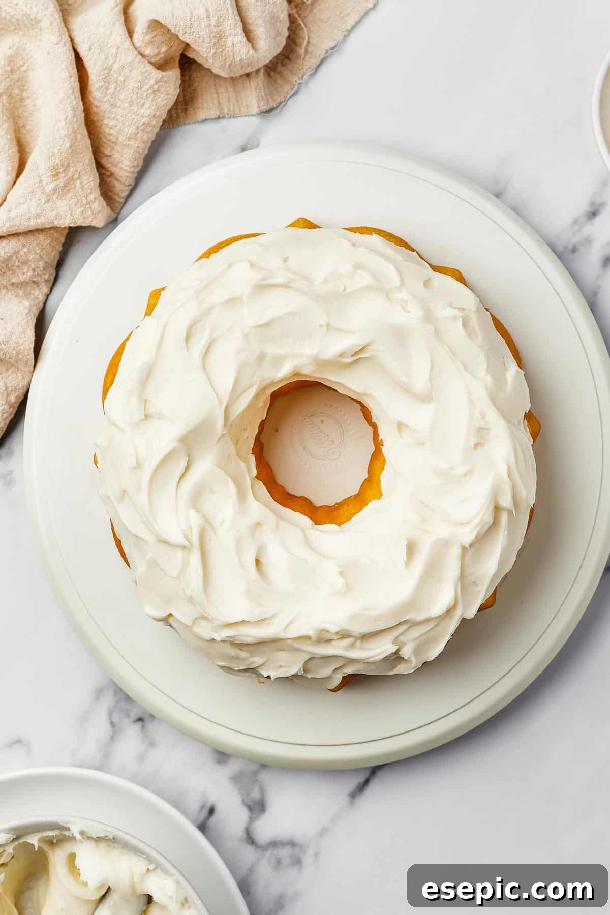 Overhead shot of a bundt cake generously iced with thick, creamy cream cheese frosting. A bowl of extra frosting and a dish of fresh lemon zest are artfully placed nearby on the table, hinting at the delightful flavors within.