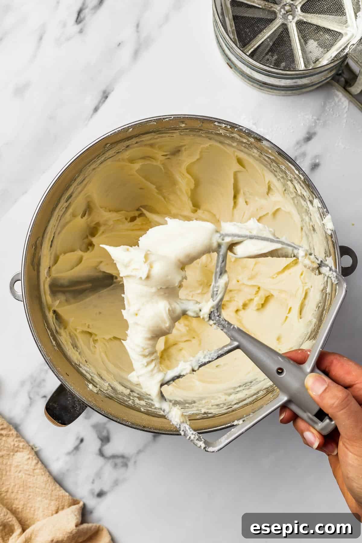 A metal mixing bowl containing whipped cream cheese frosting. A woman's hand is shown gently lifting a beater attachment out of the frosting, showcasing its fluffy consistency.