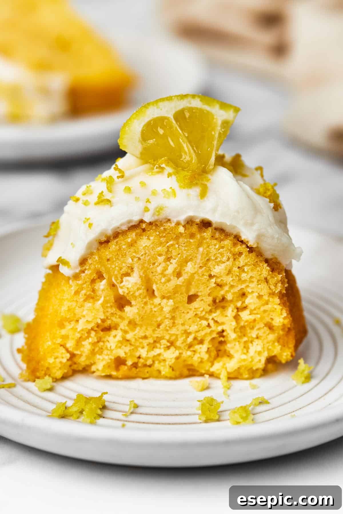 A perfectly portioned slice of frosted lemon bundt cake, elegantly garnished with a sprinkle of lemon zest and a small, fresh lemon wedge on a white plate.