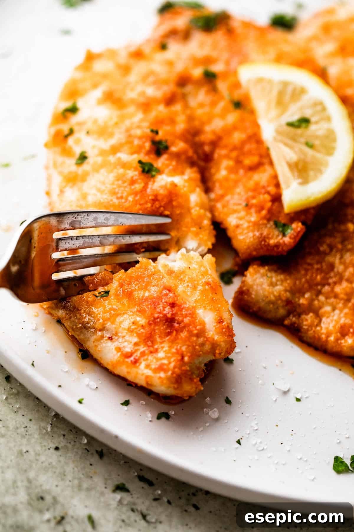A fork cuts into a crispy, golden-brown air fryer Parmesan crusted tilapia fillet, revealing its tender, white interior.
