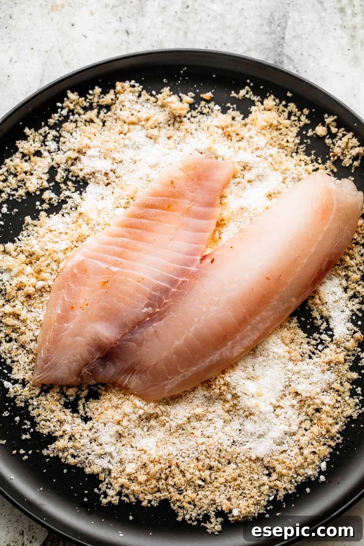 A raw tilapia fillet being pressed into a shallow dish filled with the Parmesan bread crumb mixture, ensuring full coverage.
