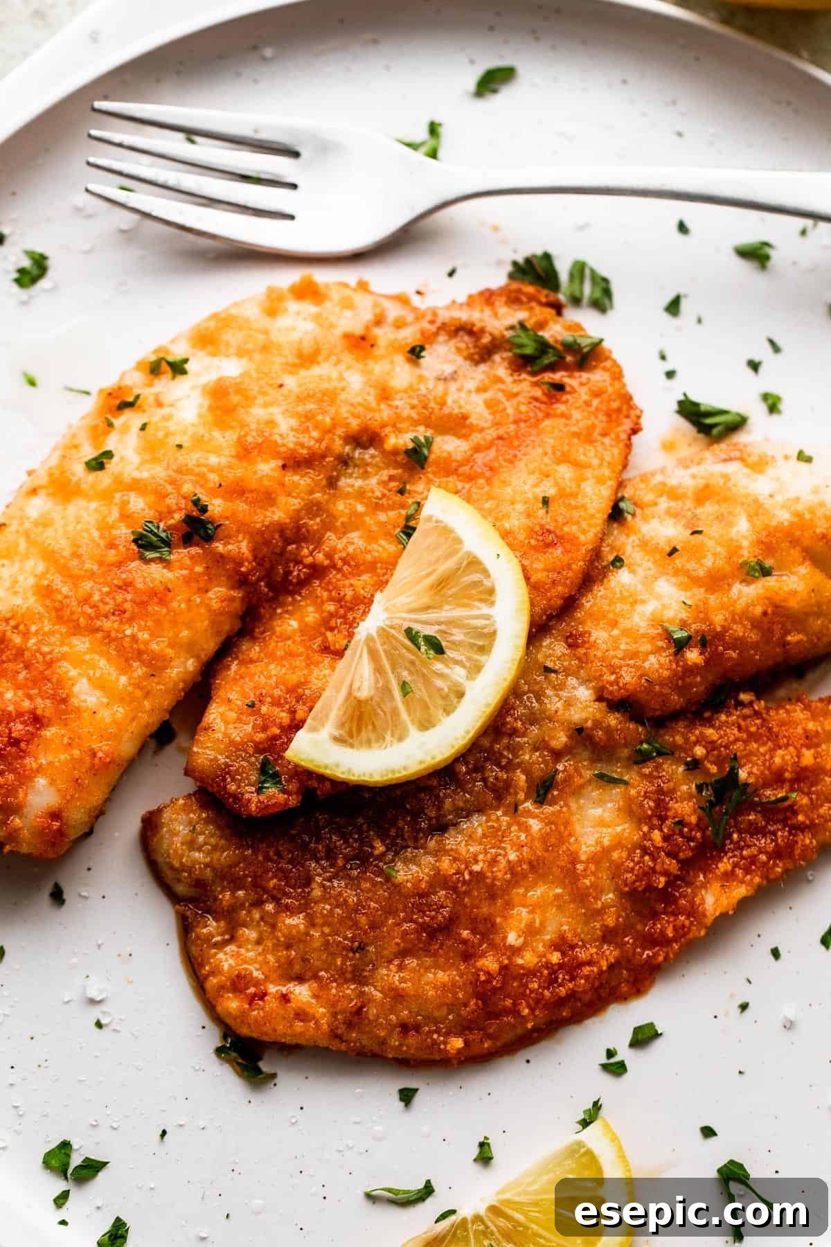 Two cooked tilapia fillets are plated beautifully on a white dish, garnished with fresh parsley and a bright lemon slice.