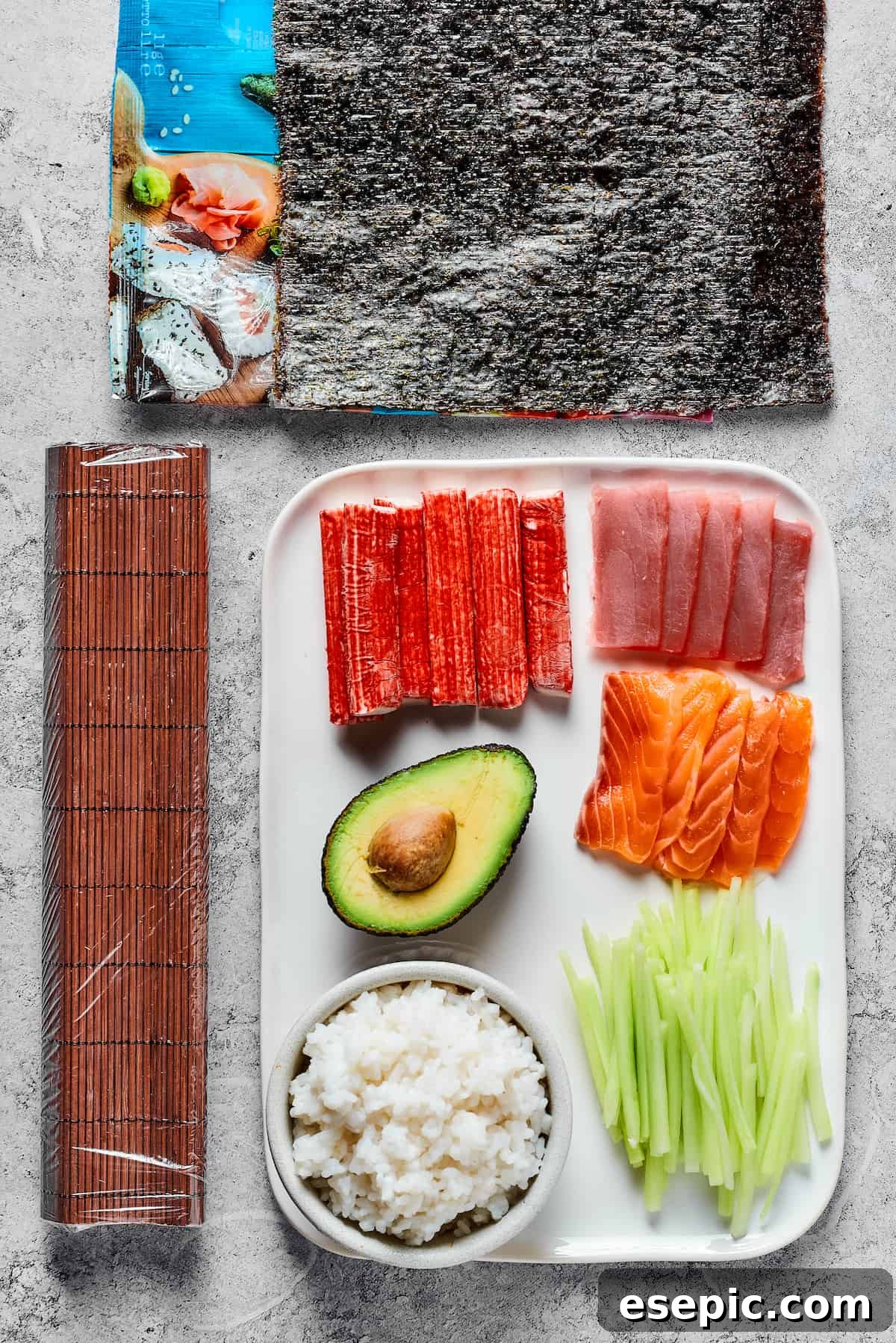 From top: Sheets of nori, a bamboo sushi mat, sticks of imitation crab, sliced raw tuna, an avocado, sliced raw salmon, prepared sushi rice, and sliced cucumber.