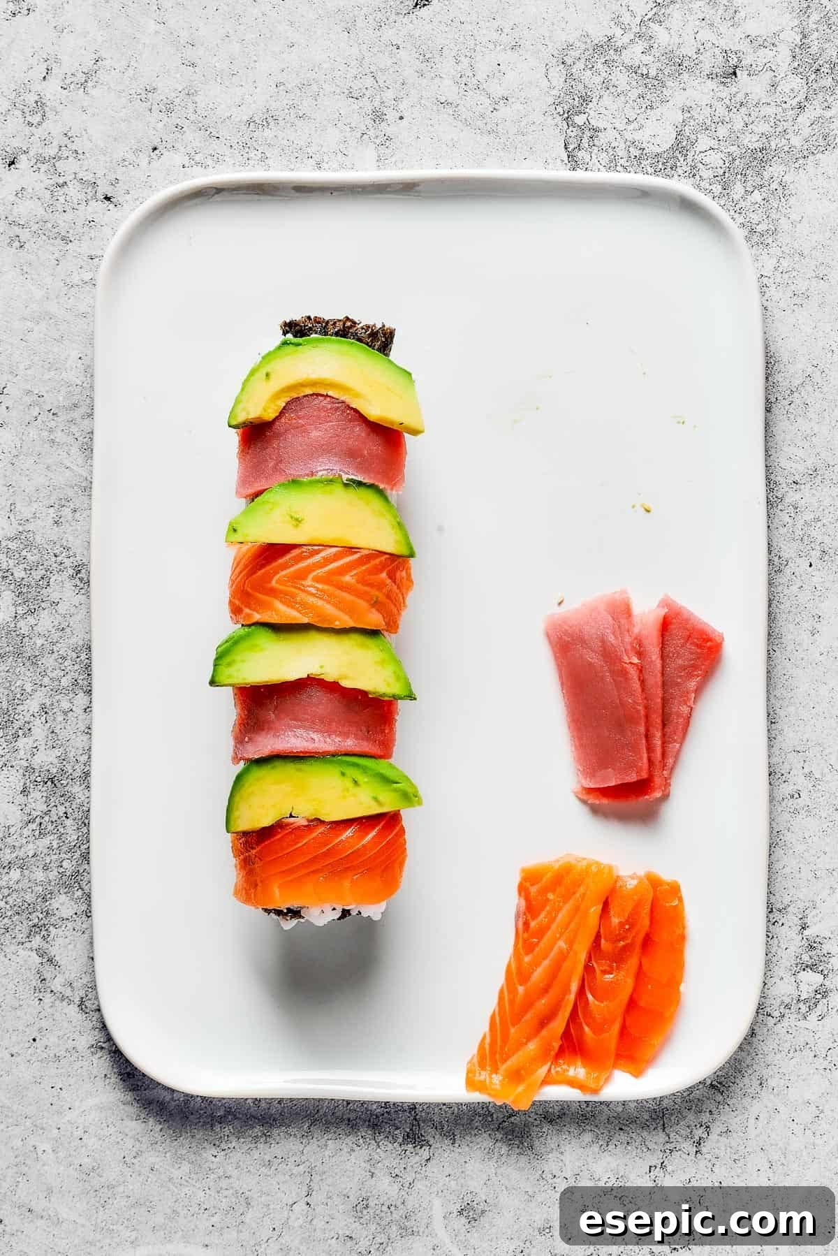 Overhead shot of a roll of sushi, topped with alternating slices of avocado, tuna, and salmon. Extra slices of tuna and salmon are on the platter.