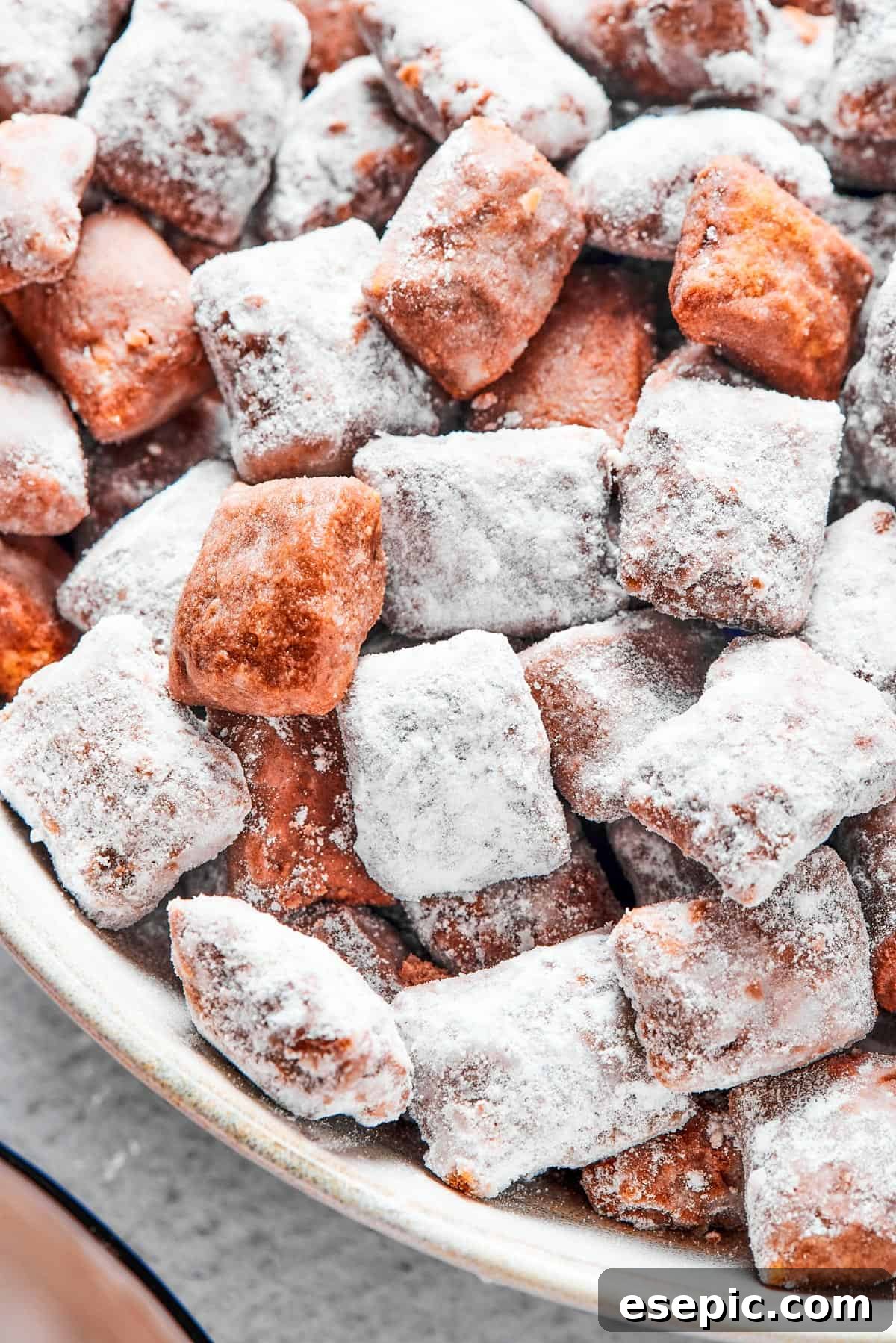 Close-up shot of puppy chow in a white bowl, to show the texture. Some pieces are fully coated in powdered sugar, while others are not.