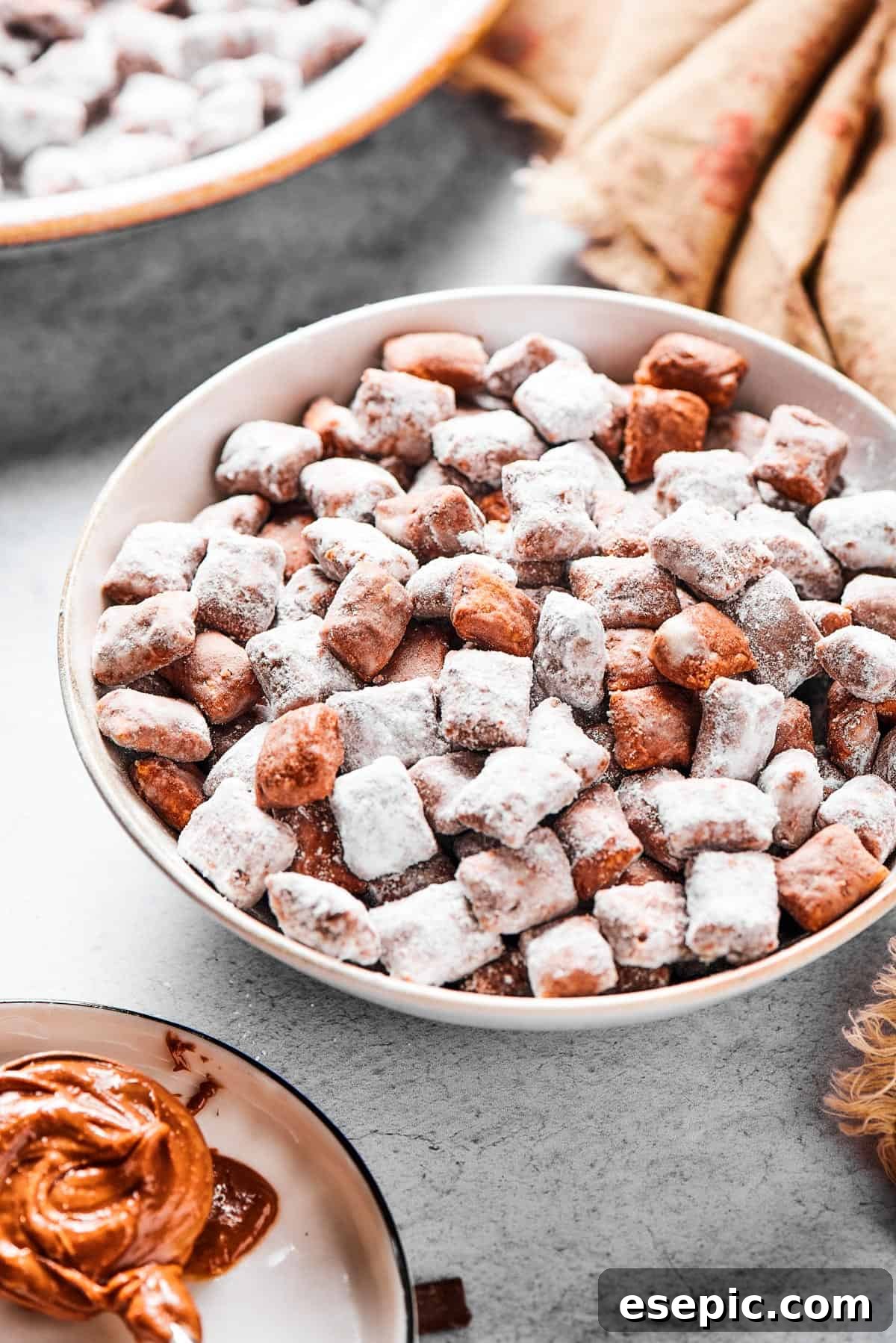 A shallow bowl filled with candy-coated cereal, near a beige napkin. Peanut butter and chocolate chunks are nearby on the table.