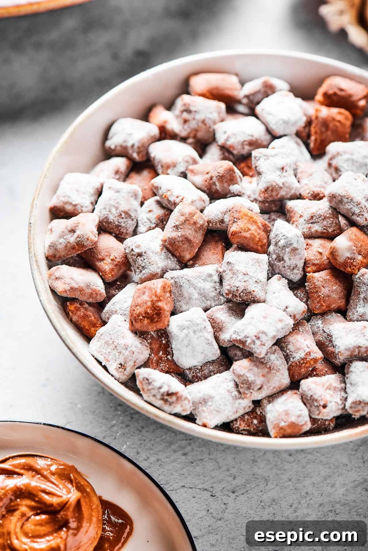 Close-up shot of puppy chow in a white bowl, to show the texture. Some pieces are fully coated in powdered sugar, while others are not.